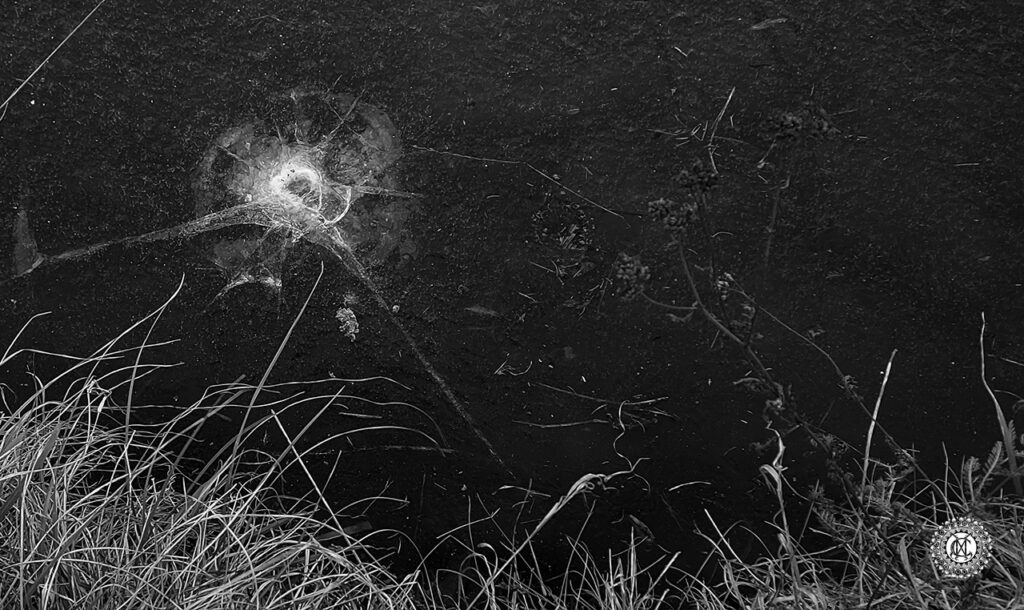 Une photo en noir et blanc, vue de dessus d’un bord de canal gelé, l’herbe de la berge est en bas de l’image, l’eau gelée est très sombre, une étoile de fissures apparaît dans la partie supérieure gauche de l’image